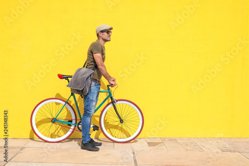 Side view of a young hipster man with a fixed bike wearing casual clothes while looking away against a yellow wall outdoors in a sunny day