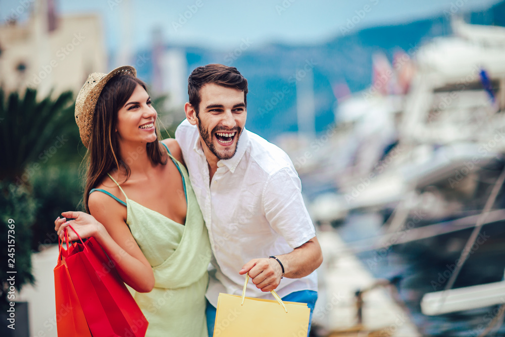 Happy young couple with shopping bags walking by the harbor of a touristic sea resort with boats on background Happy young couple with shopping bags walking by the harbor of a touristic sea resort with boats on background