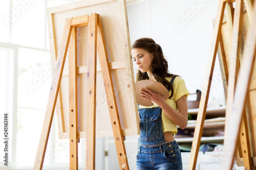 art school, creativity and people concept - student girl or young woman artist with easel and palette painting at studio