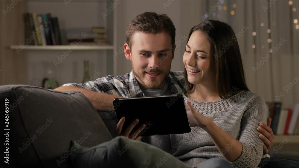 Happy couple watching online streaming videos on a tablet sitting on a couch in the night