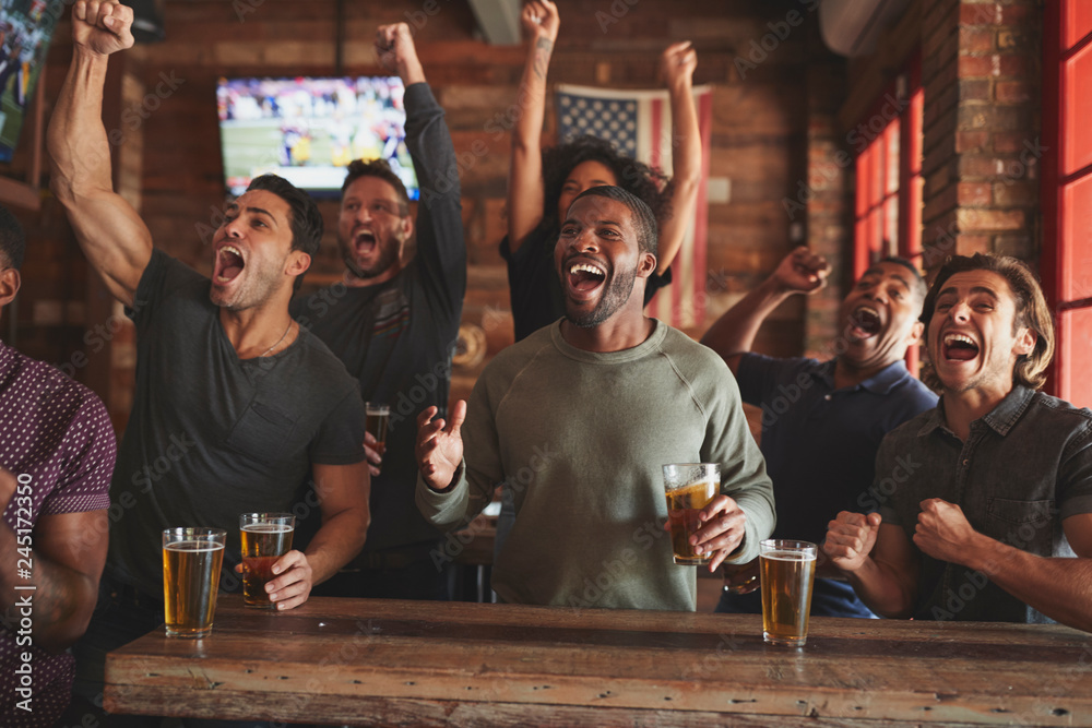 Group Of Male Friends Celebrating Whilst Watching Game On Screen In Sports Bar - obrazy, fototapety, plakaty 