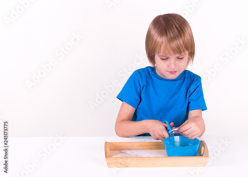 Toddler boy sits by the table with a tray of Montessori materials for a lesson of practical life on a white background, isolate. Exercise cutting with scissors.