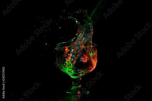 Liquid splashing out from glass with stem with vibrant colored lights on black photo