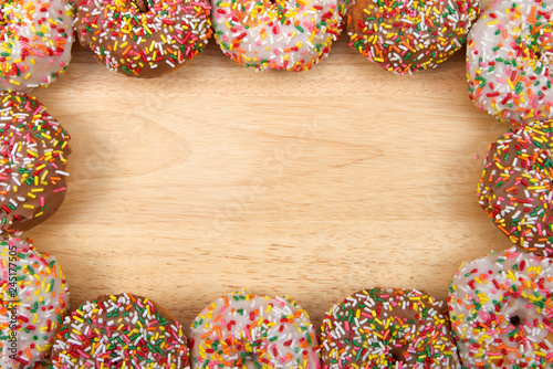 Flat lay view of many frosted donuts, chocolate and vanilla with candy sprinkles arranged in a border frame on a light wood table with copy space.