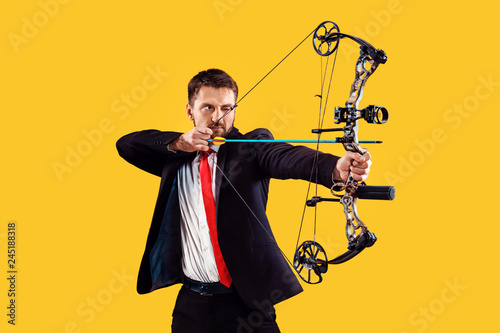 Businessman aiming at target with bow and arrow, isolated on yellow studio background. The business, goal, challenge, competition, achievement concept