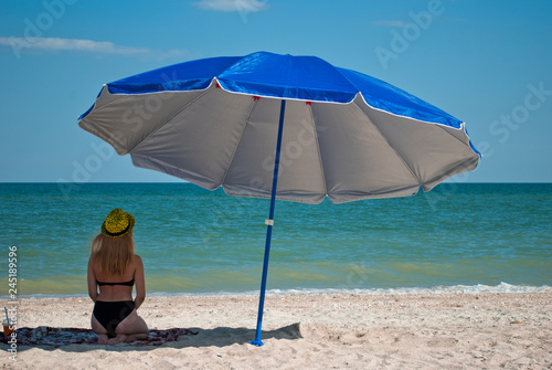 A beautiful girl sits under a beach umbrella, against the background of the sea and the sky, clouds, beach, shade, tan, hat, cap swimsuit bathing suit, summer sun weekend vacation wave foam