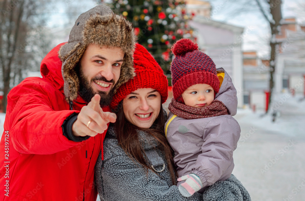 Obraz premium young energetic family. Husband and wife hug their child on the street in the middle of a snow-covered winter in a park near a tree, dressed in things of red color.