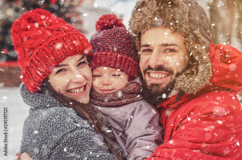 Cute family playing in the park in winter. Dad and Mom hug their child in a snow-covered winter park near a christmas tree. Enjoying spending time together. Family concept.