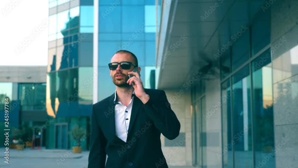 Portrait of businessman in sunglasses talking on phone and walking in street. Young man having business conversation during commuting to work. Confident guy in suit being on his way to office building Portrait of businessman in sunglasses talking on phone and walking in street. Young man having business conversation during commuting to work. Confident guy in suit being on his way to office building