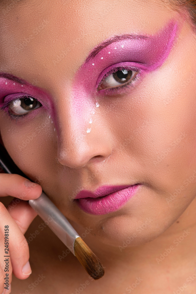 Beautiful young girl portrait, with purple and pink makeup, closeup, holding a brush close to the face, looking at the camera