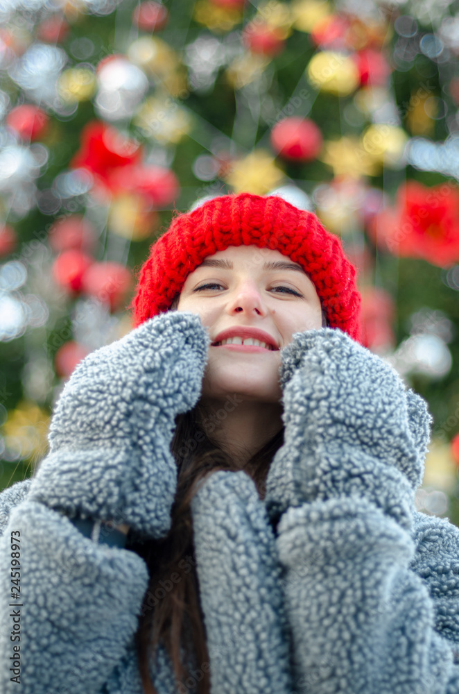 young, pretty model brunette poses for a photo in the winter near a Christmas tree in the middle of a winter city park, dressed in a red knitted hat and a warm gray coat and gloves young, pretty model brunette poses for a photo in the winter near a Christmas tree in the middle of a winter city park, dressed in a red knitted hat and a warm gray coat and gloves