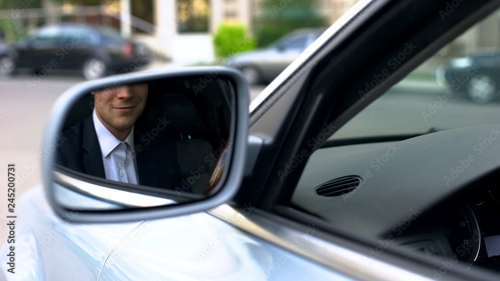 Office worker smiling in wing mirror of car, confident businessman driving
