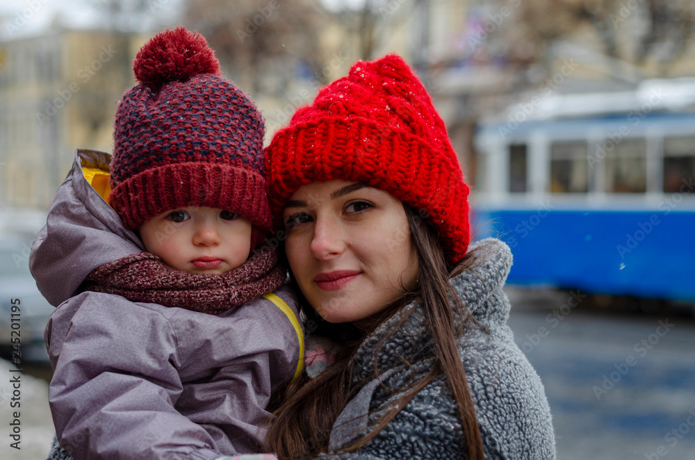 mother hugs and kisses her little baby, son or daughter, holding her in the middle of a snowy day in the city. A modern young mother wears a baby in her arms mother hugs and kisses her little baby, son or daughter, holding her in the middle of a snowy day in the city. A modern young mother wears a baby in her arms