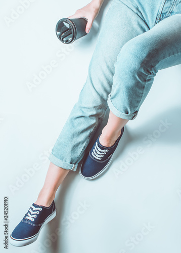 The girl in jeans and sneakers sits on a white background and holds cup of coffee. Top view. Toned photo