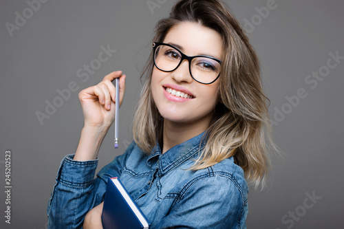 Close up portrait of a amazing woman with eyeglases and deyd hair dressed in jeans shirt and holding a notebook in one hand and pencil another smiling while looking at the camera isolated over a grey  photo