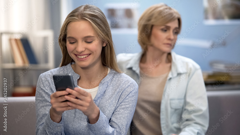Happy teenage girl texting messages in smartphone, sad mother sitting on sofa