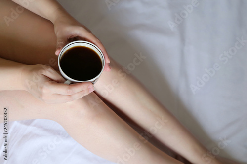 Slim girl sitting with coffee cup on the bed, top view. Coffee diet, sexy woman with naked legs and perfect smooth skin, concept of enjoying morning, relaxing at home, weekend