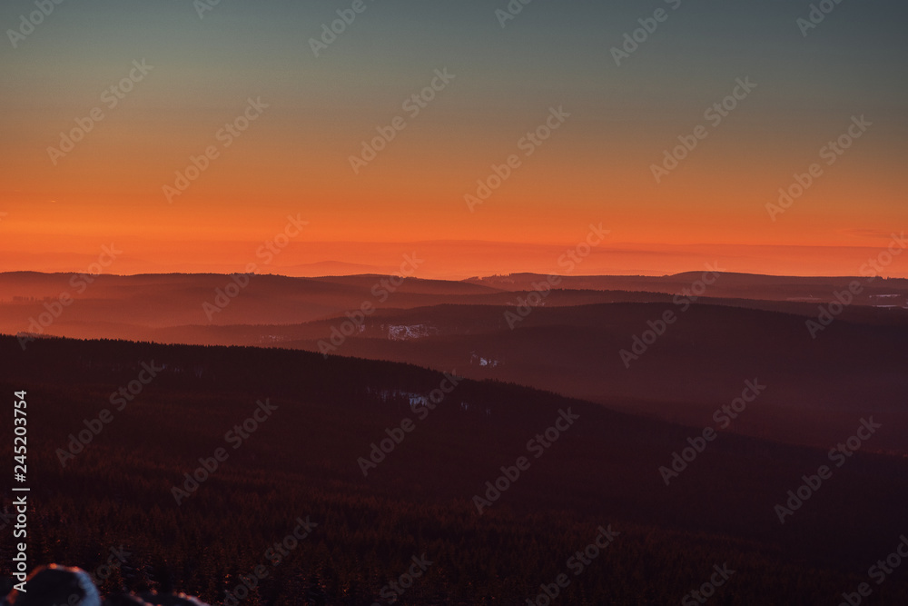 Colorful view from the mountains down to the valley with mountain silhouettes in the distance with moody sunset orange light in winter snow landscape. Torfhaus. Harz Mountains National Park in Germany