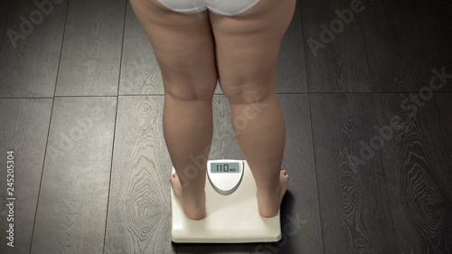 Obese female standing on bathroom scales to check body weight, fat, rear view