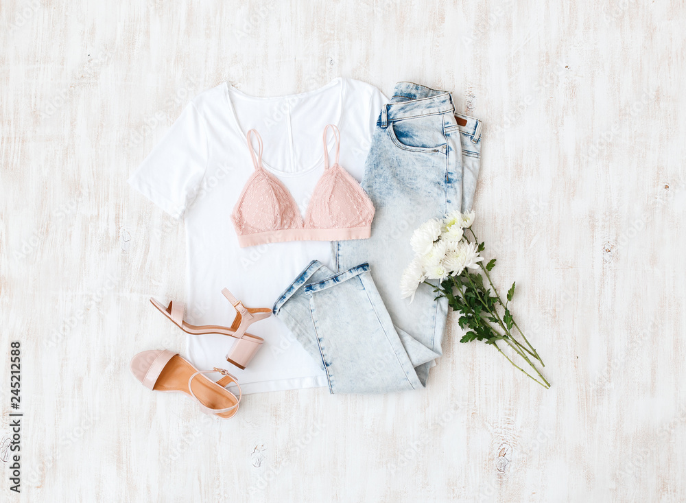 White t-shirt, blue jeans, pink lace triangle bra, pink block heeled sandals, flowers on white wooden background. Overhead view of woman's casual outfit. Simple basic women clothes. Flat lay, top view White t-shirt, blue jeans, pink lace triangle bra, pink block heeled sandals, flowers on white wooden background. Overhead view of woman's casual outfit. Simple basic women clothes. Flat lay, top view