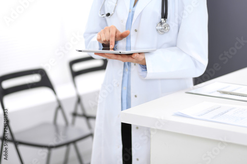 Female physician using digital tablet while standing near reception desk at clinic or emergency hospital. Unknown doctor woman at work. Medicine concept