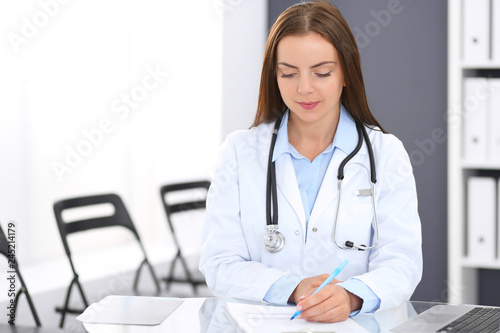 Doctor woman at work. Portrait of female physician filling up medical form while sitting at the glass desk at clinic or hospital. Medicine and healthcare concept