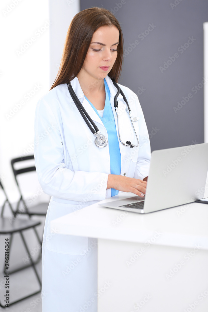 Doctor woman at work. Portrait of female physician using laptop computer while standing near reception desk at clinic or emergency hospital. Medicine and healthcare concept Doctor woman at work. Portrait of female physician using laptop computer while standing near reception desk at clinic or emergency hospital. Medicine and healthcare concept