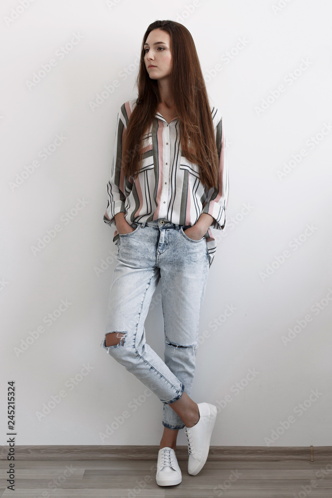 Young brunette woman, dressed in blue ripped boyfriend jeans, striped  shirt, white lace up sneakers standing in the room near white wall. Trendy  casual outfit. Simple minimalistic basic women clothes. Stock Photo