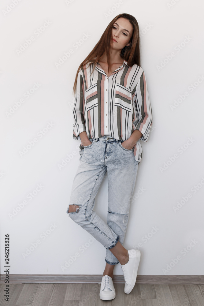 Young brunette woman, dressed in blue ripped boyfriend jeans, striped  shirt, white lace up sneakers standing in the room near white wall. Trendy  casual outfit. Simple minimalistic basic women clothes. Stock-Foto