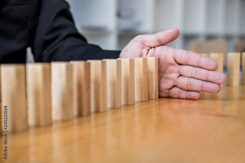 Wooden game strategy, Businessman hand stopping falling wooden dominoes effect from continuous toppled or risk, strategy and successful intervention concept for business Wooden game strategy, Businessman hand stopping falling wooden dominoes effect from continuous toppled or risk, strategy and successful intervention concept for business
