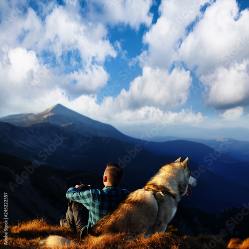Alone tourist sitting with Alaskan Malamute dog in mountains. Man with his dog on mountain top. Hiker with dog looking at beautiful view in mountains © Serhii