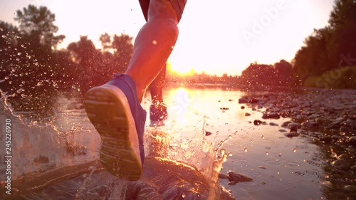 SUPER SLOW MOTION, CLOSE UP, SUN FLARE: Golden evening sunbeams shine on athletic young man jogging along the tranquil river. Picturesque serene nature surrounds sportsman running in the shallow water photo