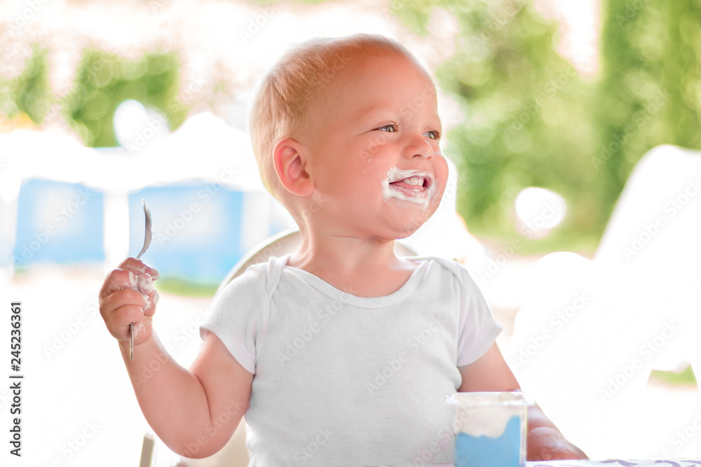 Smiling little blonde toddler boy eating delicious yogurt sitting outdoor Smiling little blonde toddler boy eating delicious yogurt sitting outdoor