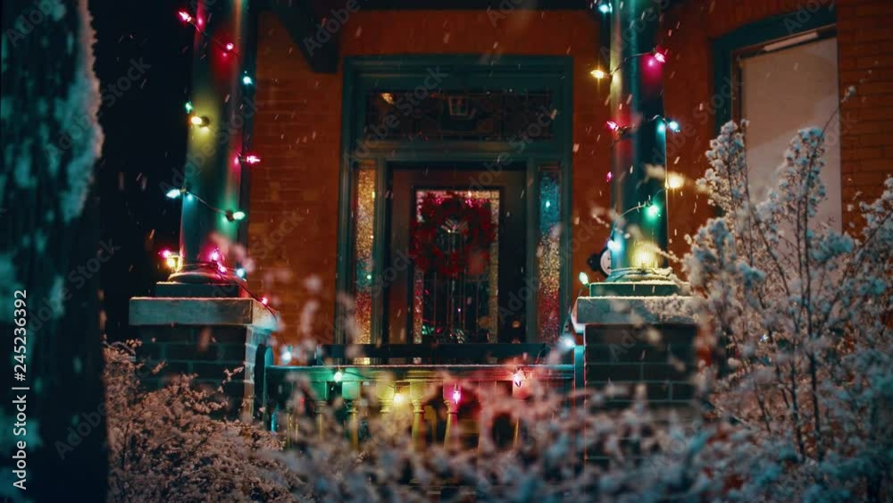 Snow falling outside of house with Christmas lights and a wreath on the front door. 