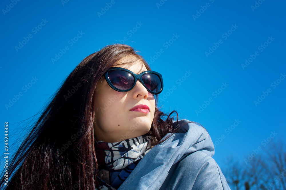 Cute young gilr wearing dark sunglasses posing outdoors over blue sky during early spring time. Cute young gilr wearing dark sunglasses posing outdoors over blue sky during early spring time.