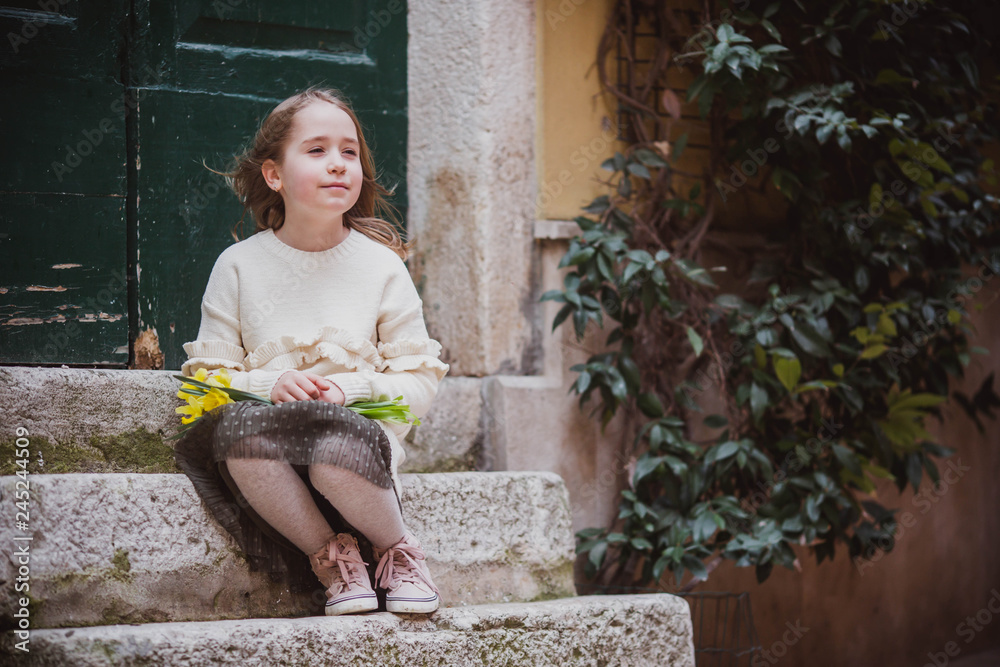 Adorable little girl in trend clothes sits near old door in old town in sunny spring day