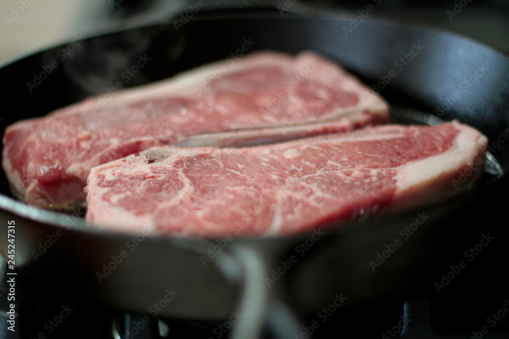 New York strip steaks cooking in a cast iron pan on a natural gas stove top.

