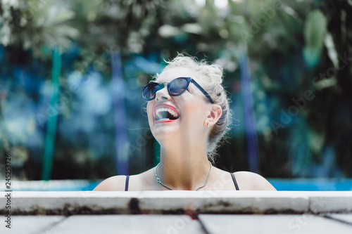 Blurred background of aromatic sticks with young beautiful laughing blonde woman in sunglasses with a good figure with red lips make-up posing in a pool of blue water. Outdoor portrait close up. 