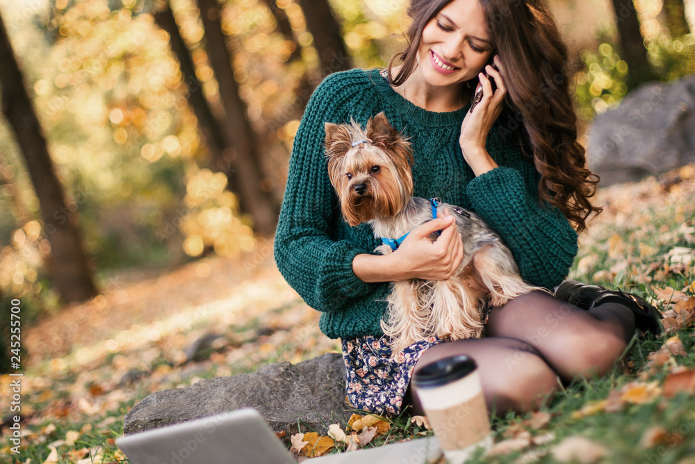Woman working on laptop and talking on the phone in the park with dog in her arms.