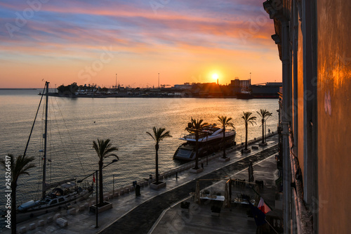 The sun sets over the harbor port as boats dock along the waterfront promenade in the coastal city of Brindisi, Italy, in the Puglia region