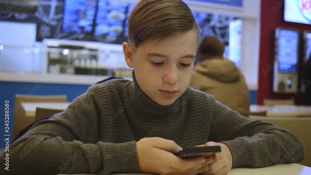 A boy sitting at a table in a cafe communicates with friends and plays on a smartphone A boy sitting at a table in a cafe communicates with friends and plays on a smartphone