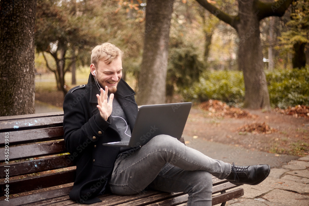 one young laughing man, sitting on bench in public park, using laptop, talking over Internet, video chat or call, could be with family, friend. gesturing one young laughing man, sitting on bench in public park, using laptop, talking over Internet, video chat or call, could be with family, friend. gesturing