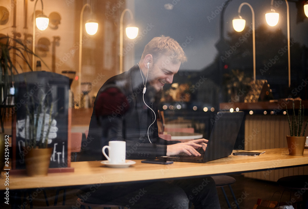 one young smiling man, 20-29 years, sitting in coffee shop and using, typing on keyboard of his laptop. Shoot thought window outside (with reflections from outside).