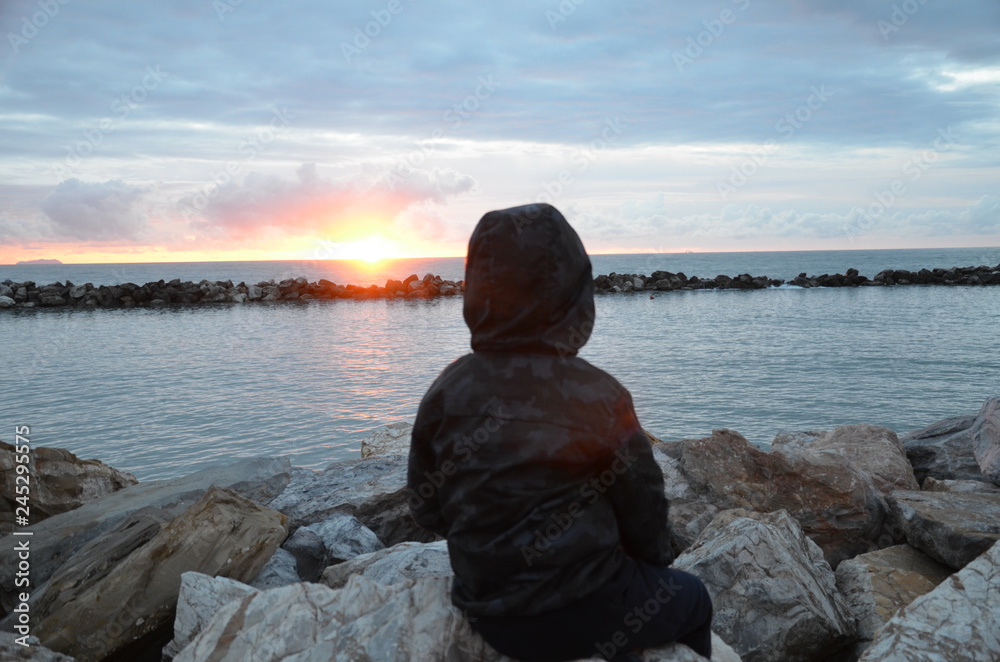 Young boy sitting on a rock by the sea and watching the sunset