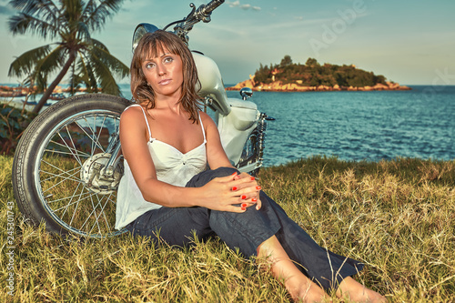 Young Woman sitting on the grass at thevintage custom motorbike on the tropical coast background. Fashion female biker concept. photo