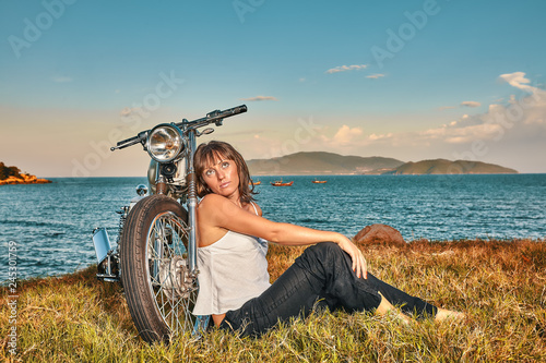 Young Woman sitting on the grass at thevintage custom motorbike on the tropical coast background. Fashion female biker concept. photo