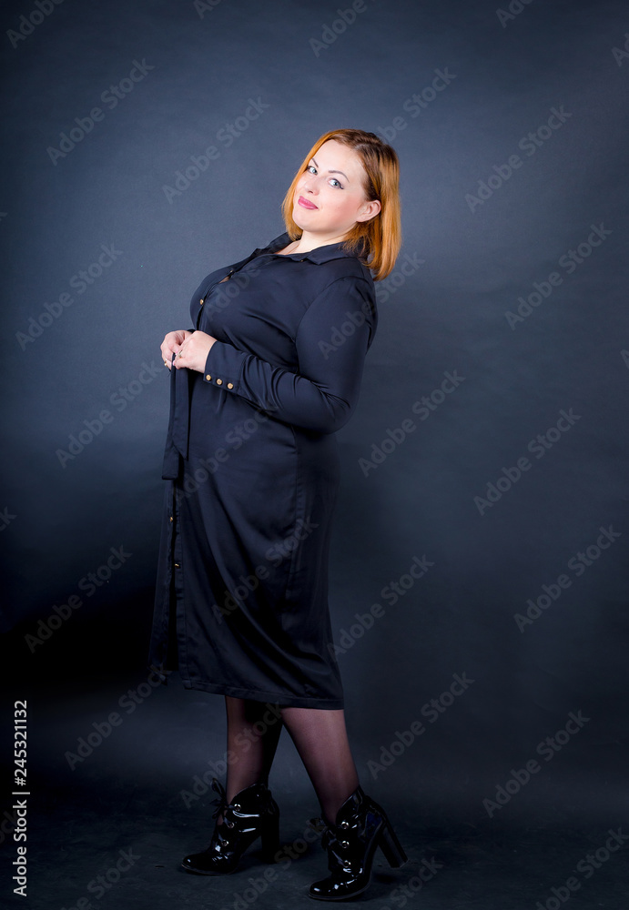 Beautiful chubby girl in a black dress on a dark background. Self confident plus size woman in glasses and black dress posing in frame Stock Photo Adobe Stock