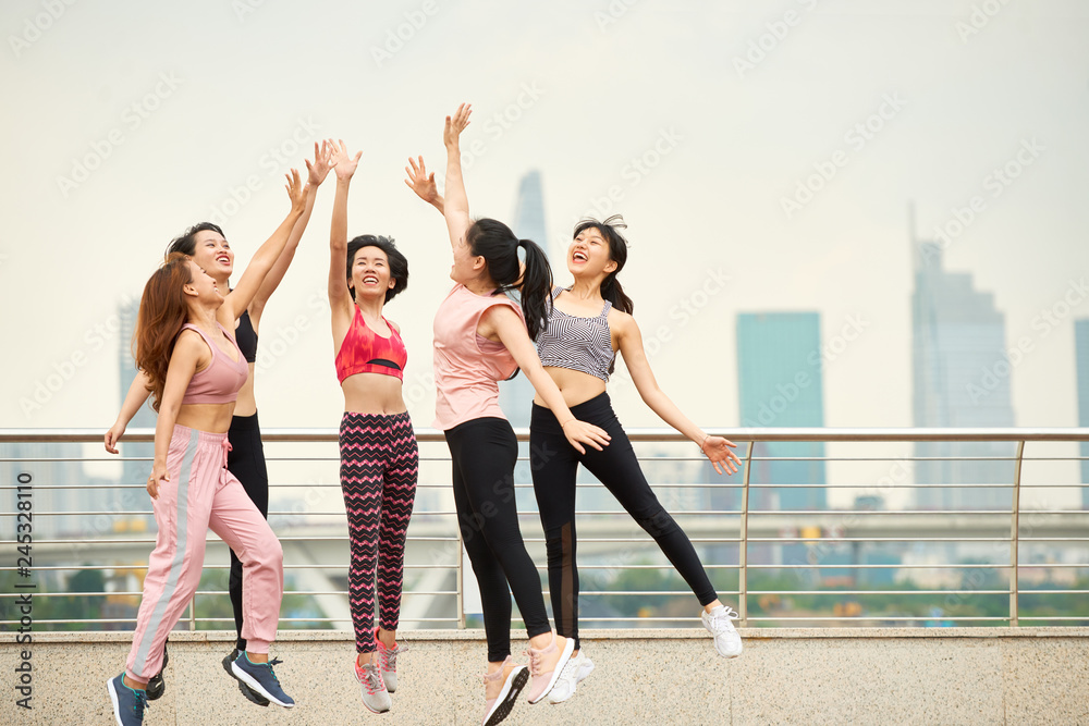 Fototapeta premium Group of laughing Asian woman in sportswear jumping and giving high five against cityscape 