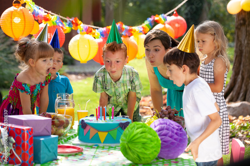 Redhead boy, his mother and friends blowing candles on birthday cake during garden party for children