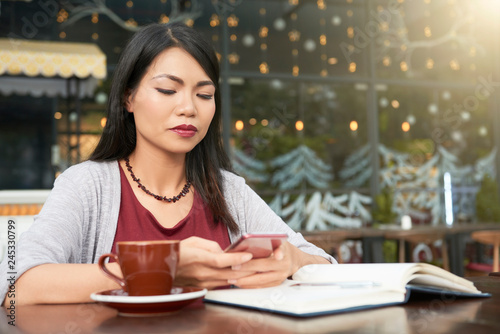 Young attractive woman sitting at the table with notepad and cup of coffee and using her smartphone at outdoor cafe